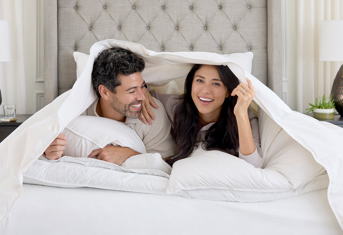 A smiling couple lies under a white duvet on a bed in a bright bedroom. The man wears a light beige shirt, and the woman wears a light pink top.