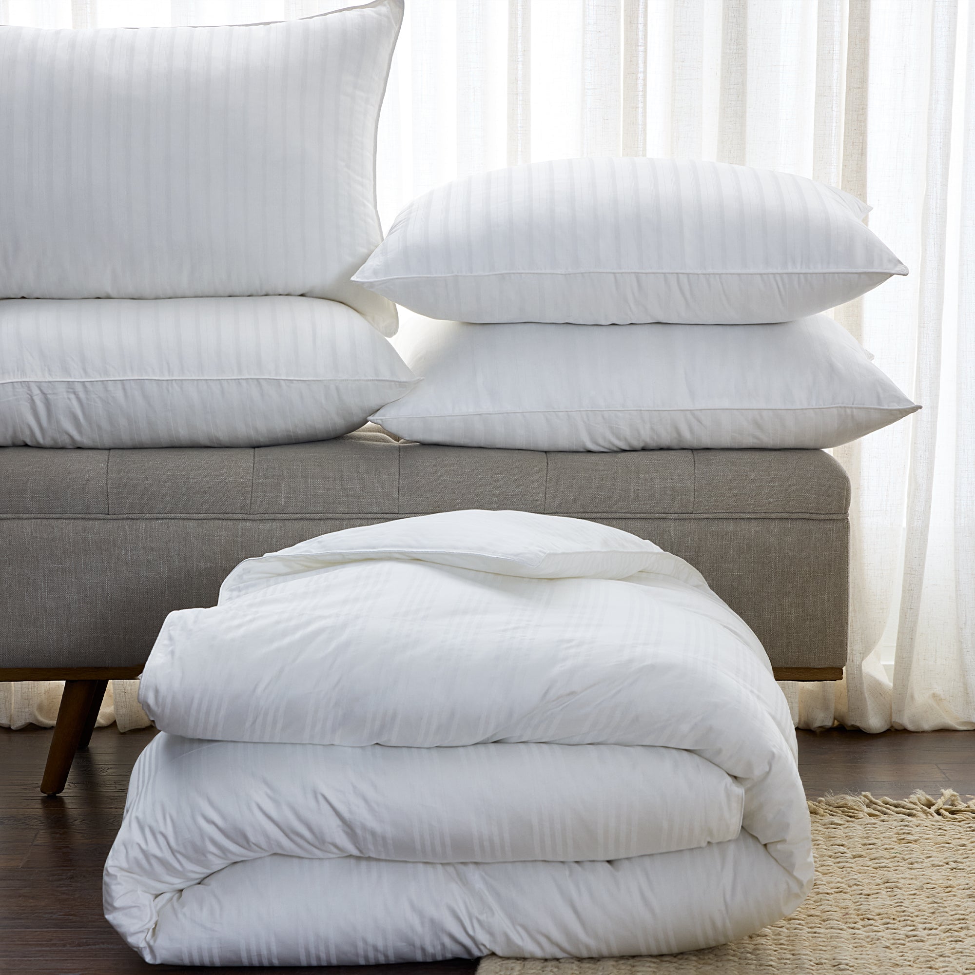 Stack of white pillows and a folded white comforter on a gray sofa with a neutral background.