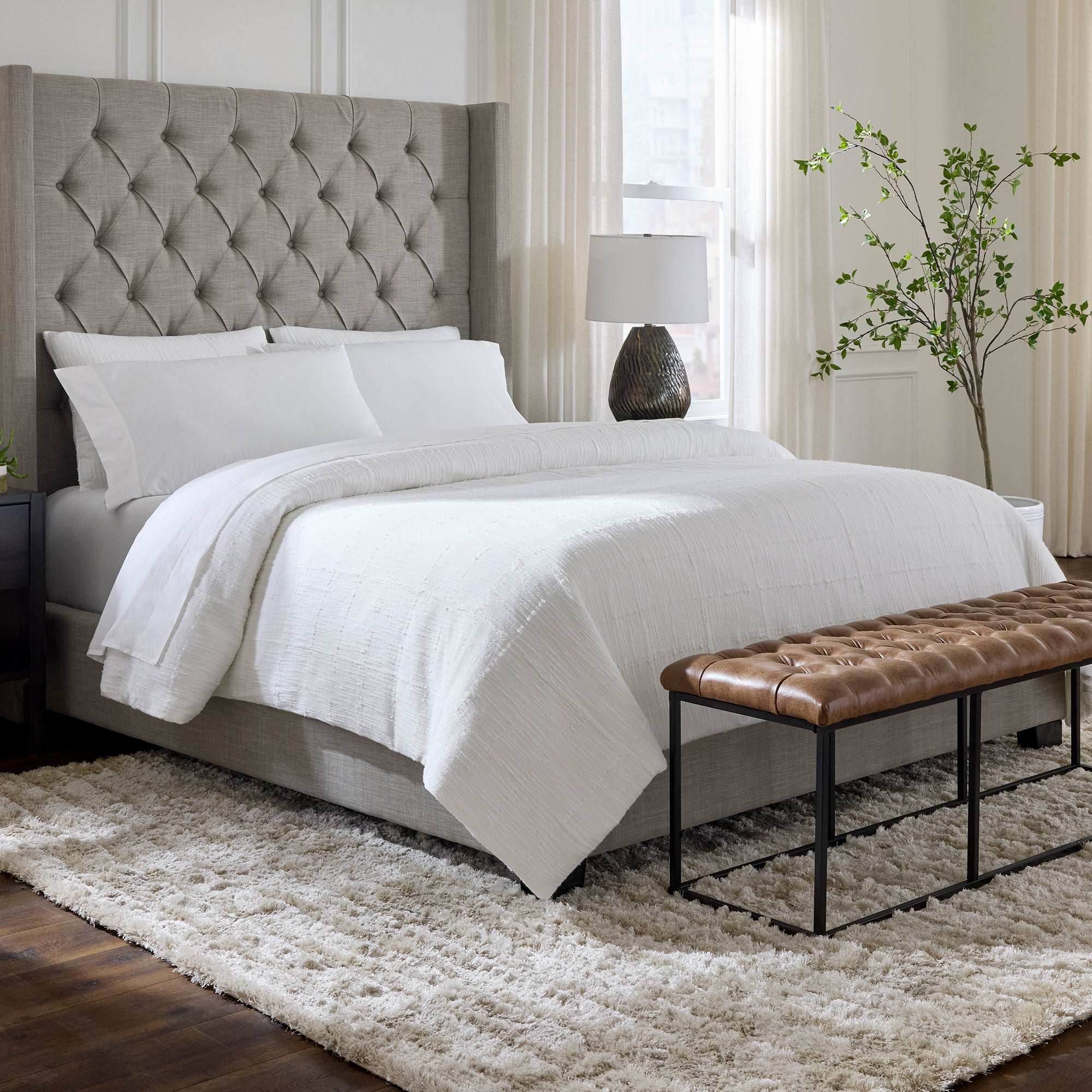 Neatly made bed with white bedding in a well-lit bedroom with a brown bench and lamp.