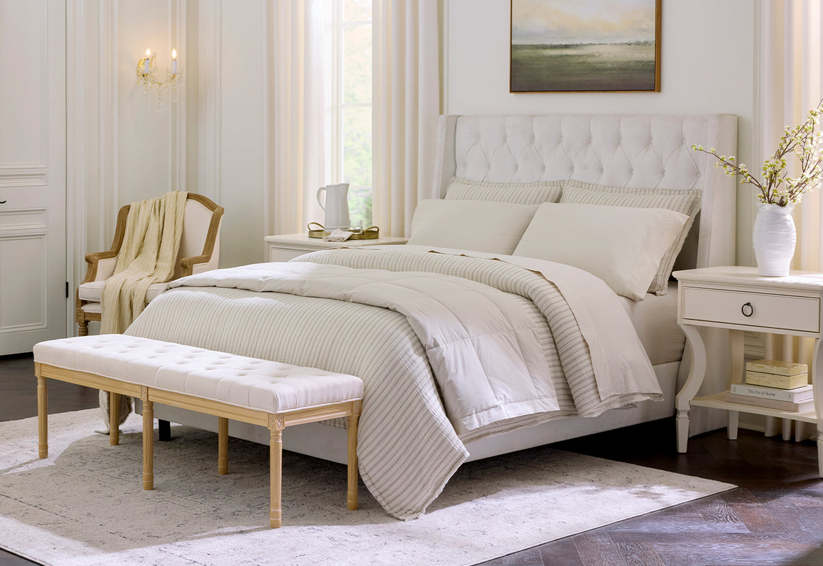 A beautifully styled bedroom featuring a cream tufted bed frame, striped cream and beige bedding, a matching tufted bench, and a light wood chair on a patterned area rug.