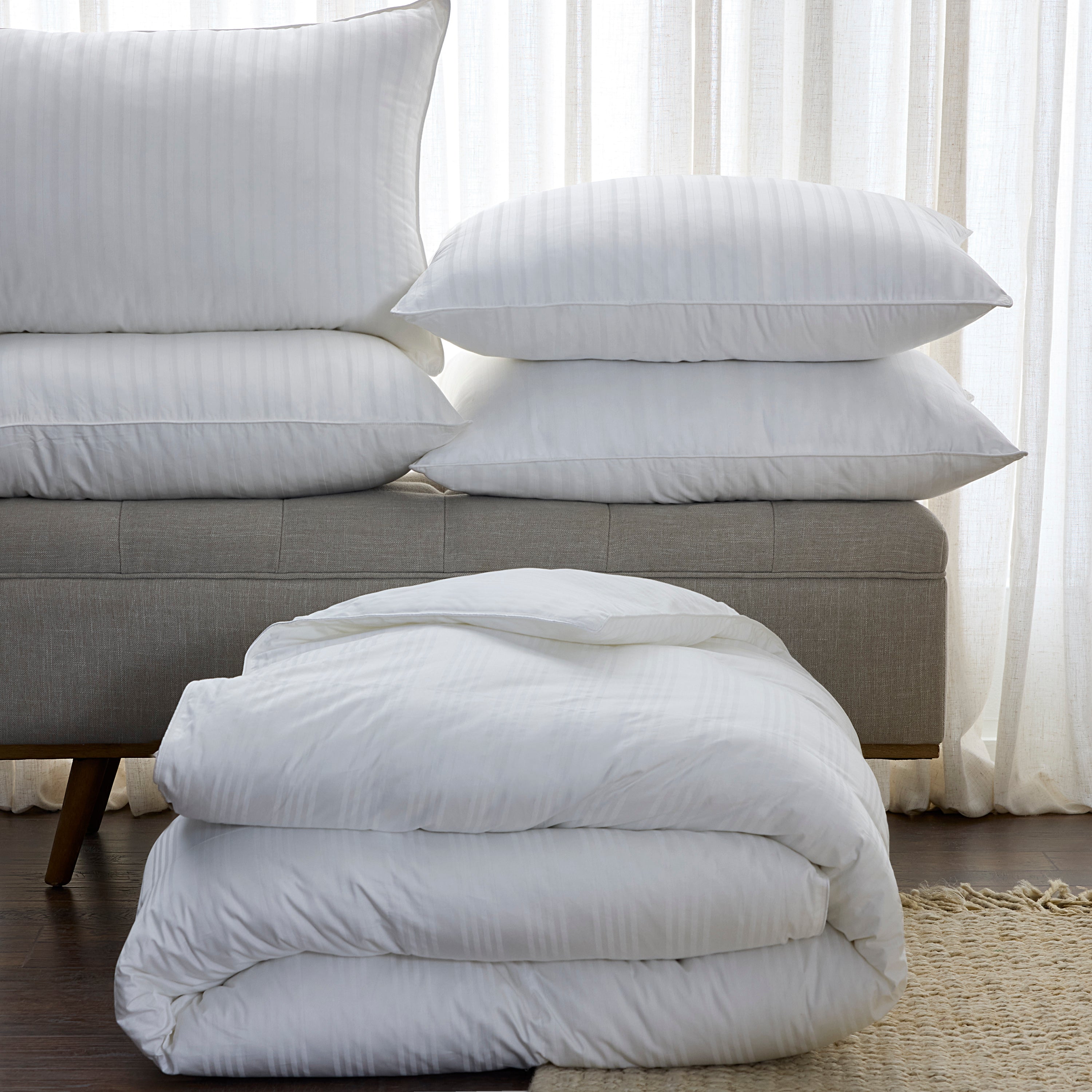 Stack of white pillows and duvet on a gray sofa with white curtains in the background