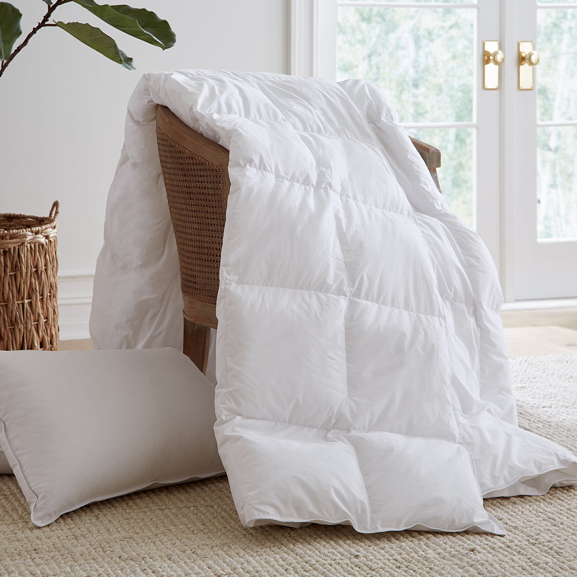 White comforter draped over a wicker chair in a bright room with large windows.